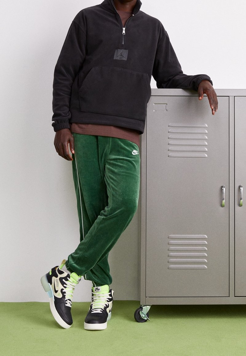 Felpa nera con zip, pantaloni di velluto verde e sneakers nere e bianche con accenti al neon, posizionati accanto a un armadietto argentato.