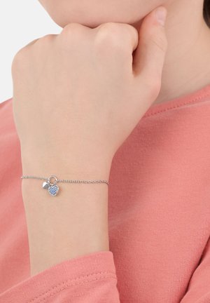 Pulsera de cadena de plata con un charm de corazón con textura sutil y un acento de corazón más pequeño. Llevada en una mano contra una blusa rosa.