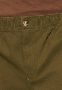 Παντελόνι ελιάς με ελαστική μέση και κούμπωμα με κουμπί, φορεμένο με καφέ κορυφή μέσα στην μέση.