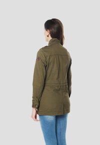 Olijfgroene canvas jas met een getailleerde taille, hoge kraag en geborduurde accenten op de schouders. Manchetten met knopen en detail in het rugpand.