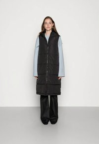 Νεαρή γυναίκα που φοράει ένα μακρύ μαύρο γιλέκο με κουκούλα πάνω από ένα ανοιχτό μπλε πουλόβερ και μαύρες φαρδιές παντελόνες, στέκεται σε ένα γκρίζο δάπεδο.