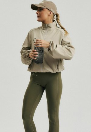 Femme portant une veste kaki, des leggings vert olive et une casquette beige, tenant une bouteille d'eau noire, regardant sur le côté.