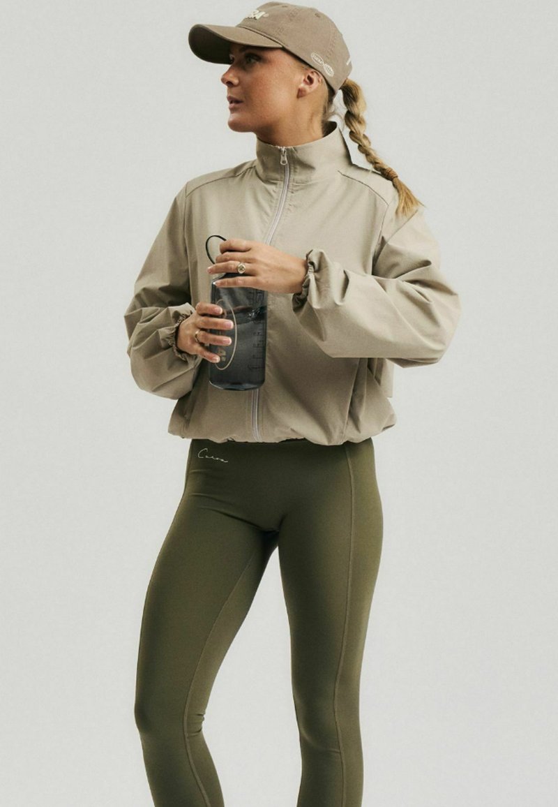 Femme portant une veste kaki, des leggings vert olive et une casquette beige, tenant une bouteille d'eau noire, regardant sur le côté.