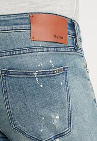 Jeans de mezclilla azul deslavada con manchas de pintura blanca. Presenta un parche de cuero marrón en la cintura con la marca "tigha" y un acento de hardware plateado.