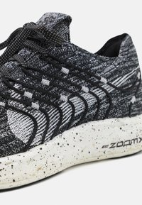 Zwart-wit gestreepte sportschoen met een getextureerde gebreide bovenkant, ronde veters en een gespikkelde witte tussenzool. ZoomX-logo zichtbaar.