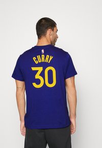 Camiseta de la NBA de color púrpura con "CURRY" en letras amarillas encima del número "30". Diseño de cuello redondo, hecha de tejido suave y de mangas cortas.
