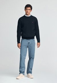 Zwarte kabelgebreide trui met geribde manchetten en zoom, voorzien van een klein rood logo. Gecombineerd met lichtblauwe denim jeans en beige sneakers.