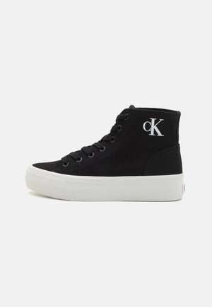 Czarny sneaker za kostkę z białą grubą podeszwą, czarnymi sznurowadłami i białym logo "cK" na części przy kostce, pokazany z boku.