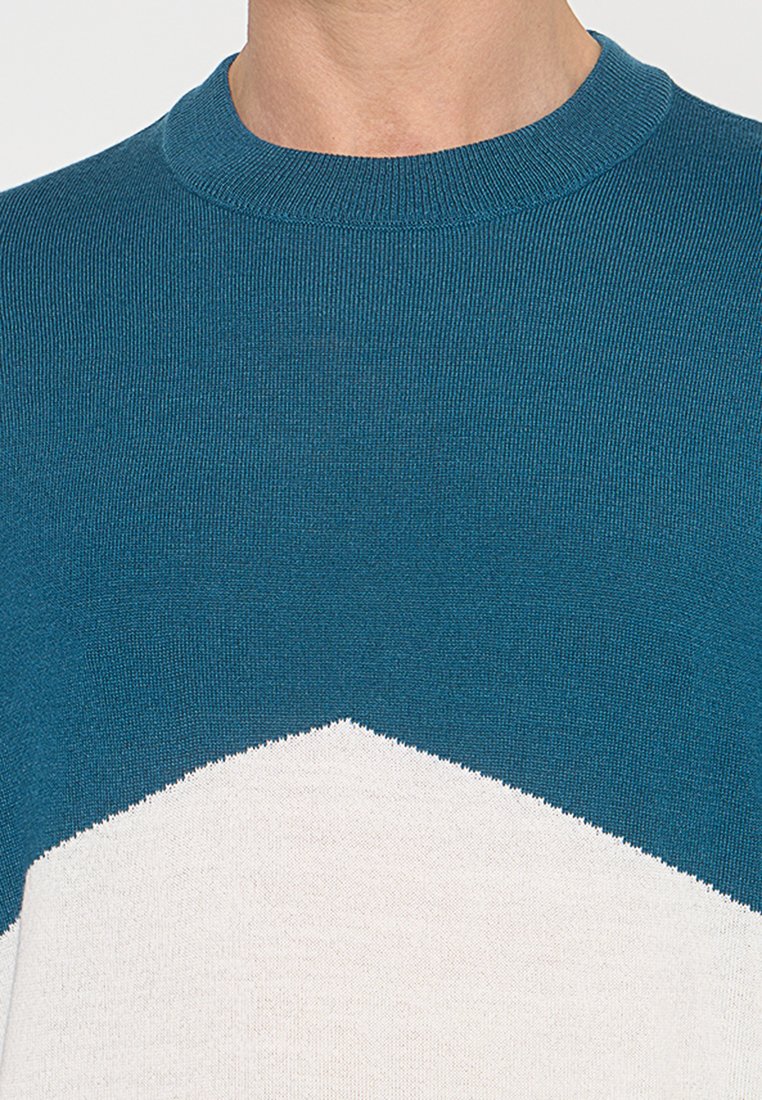 Krupan plan osobe koja nosi tealo i bijeli pleteni pulover s okruglim izrezom i chevron uzorkom na prsima.