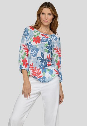 Frau trägt eine langärmelige Bluse mit roten, blauen und grünen Blumen, kombiniert mit weißen Hosen, steht vor einem einfarbigen grauen Hintergrund.