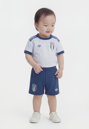 Tout-petit debout, portant un maillot et un short de football italien blanc et bleu avec des baskets blanches assorties, regardant légèrement sur le côté.