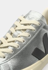 Sneaker argento con finitura texture, lacci crema, dettaglio logo nero e suola spessa color crema. Vista ravvicinata che mette in evidenza gli elementi di design.
