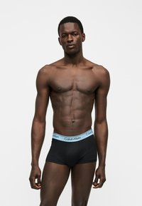 Atletisk mørk hudfarvet mand står, iført sorte Calvin Klein boxershorts med lyseblåt taljebånd, mod en ensfarvet hvid baggrund.