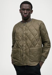 Chaqueta acolchada de color verde oliva con cuello redondo, dos bolsillos frontales y puños acanalados. Material suave y ligero con un patrón de diamante.