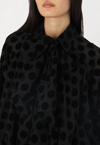 Black sheer blouse with large, textured polka dots. Features a bow tie collar and flowing ruffles along the front, enhancing its layered design.
