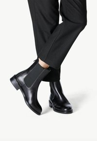 Botas de cuero negras hasta el tobillo con paneles laterales elásticos, punta redondeada y un tacón bajo cuadrado, combinadas con pantalones oscuros.