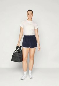 Sort duffel bag, lyserød t-shirt, marineblå shorts med hvid kant, hvide sokker og sneakers. Shortsene og tasken har en blød tekstur.