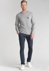 Maglietta grigia a maniche lunghe con scollatura rotonda e un piccolo logo; abbinata a pantaloni slim-fit blu navy e sneaker bianche. Dettagli di design semplici.