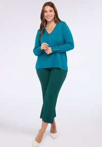 Uśmiechnięta kobieta stoi w turkusowym swetrze w serek z złotymi guzikami na rękawach, ciemnozielonych spodniach w kant, oraz białych szpilkach.