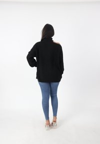 Maglione nero oversize a maglia con colletto alto e dettagli sulle spalle scoperte, abbinato a jeans blu aderenti e tacchi trasparenti.