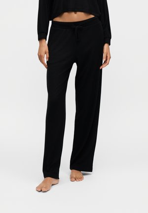 Persoon draagt zwarte, loszittende broek met een trekkoord in de taille en een zwarte cropped top met lange mouwen, staat op blote voeten.