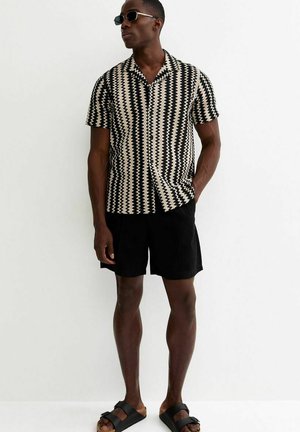 Mężczyzna ubrany w czarno-białą koszulę z krótkim rękawem w zygzakowaty wzór, czarne szorty, czarne sandały i okulary przeciwsłoneczne stoi na tle białej ściany.