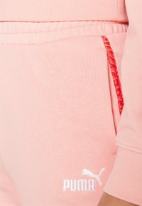 Rosa bomullbukser har en hvit brodert logo, elastisk midje og en rød mønstret aksent på lommen.