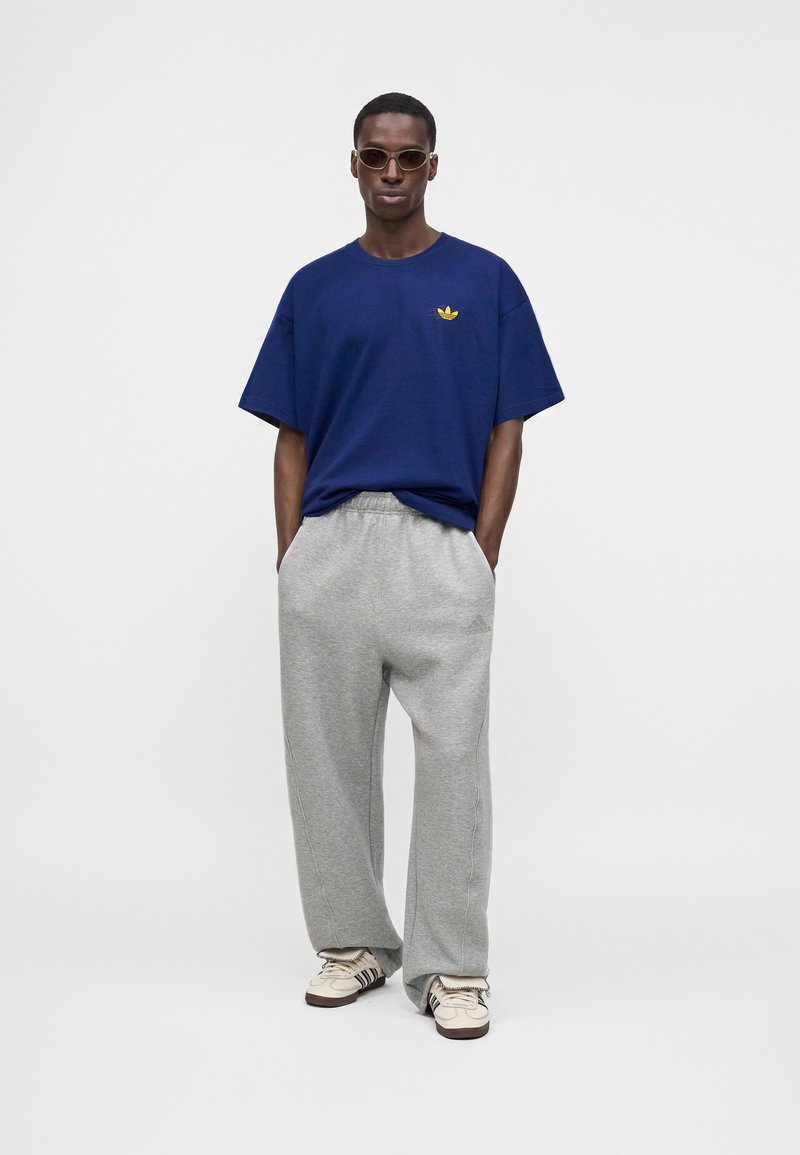 Hombre con camiseta azul oscuro con logo amarillo, pantalones deportivos grises, zapatillas blancas y gafas de sol, de pie frente a un fondo claro y liso.