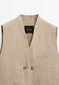 Beige kordflöjelväst med V-hals, dubbel knappstängning och en bröstficka. Texturerat tyg med vertikala ränder. Märkesetikett synlig.