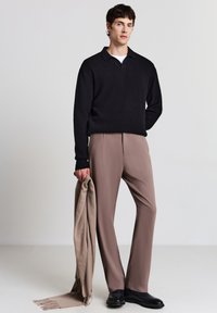 Schwarzer gerippter Strick-Polo-Pullover, beiges Tailoring-Hose, hält einen hellbraunen Schal, schwarze Lederschuhe, vor einem schlichten hellen Hintergrund.