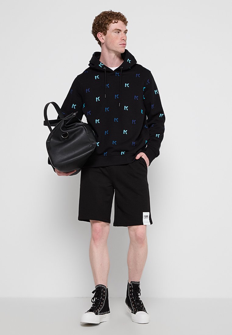 Sweat à capuche noir avec un logo "K" multicolore, associé à un short noir et des baskets montantes noires. Tient un grand sac noir.