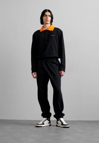 Svart langermet polo med oransje krage, kombinert med svarte joggebukser og svart-hvite sneakers, står på en grå gulv.
