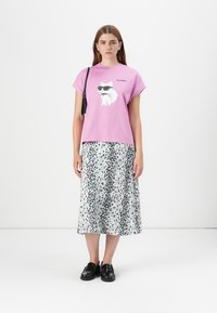 Le mannequin se tient debout portant un T-shirt rose avec un chat en cartoon portant des lunettes de soleil, associé à une longue jupe à motifs et des chaussures noires, sur un fond uni.