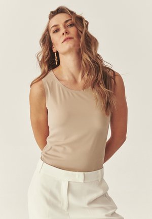 Blondynka o falowanych włosach, ubrana w beżowy bezrękawnik, białe spodnie z wysokim stanem i długie, skręcone złote kolczyki, stoi na jednolitym tle.