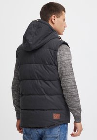Chaleco negro sin mangas acolchado con capucha, con un diseño acolchado y un parche de logo marrón en la parte posterior, usado sobre un suéter gris.