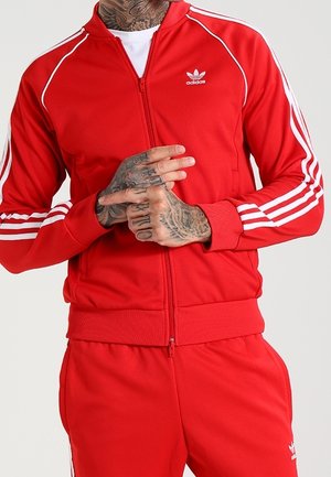 Mand iført rød Adidas træningsdragt med hvide striber og logo, der viser tatoverede hænder og nakke, stående foran en ensfarvet baggrund.