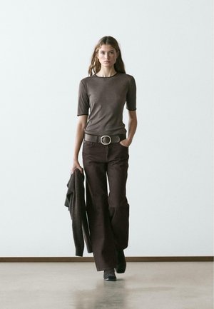 T-shirt marron ajusté à manches courtes, associé à un pantalon marron évasé, une large ceinture et un cardigan sombre drapé sur un bras.