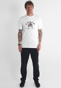 T-shirt blanc à manches courtes avec un imprimé graphique bleu et jaune. Porté avec un pantalon foncé et des baskets noires, posé devant un fond uni.