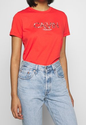 Person wearing a bright red Calvin Klein t-shirt with leopard print letters, tucked into high-waisted light blue jeans.