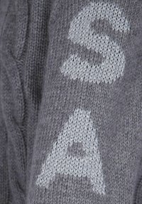 Tessuto grigio lavorato a maglia con filo metallico grigio chiaro che forma la lettera "S" sopra la lettera "A" su una superficie testurizzata.