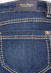 Jeans de mezclilla azul con una etiqueta que dice "Socax Denim Established 1996", con costuras beige en contraste y un pequeño acento metálico.