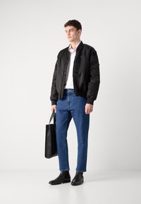 Chaqueta bomber negra sobre una camiseta blanca, combinada con jeans de mezclilla azules y botas chelsea negras. Sosteniendo un bolso tote negro. Fondo de estudio.