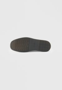 Suela de zapato de goma negra con un patrón ondulado, presentando un diseño suave y ligeramente elevado. Logotipo de la marca "Calvin Klein" grabado en la superficie.