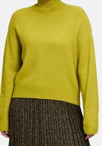 Limettengrüner Strickpullover mit hohem, geripptem Kragen und zulaufenden Ärmeln. Kombiniert mit einem plissierten Rock mit Leopardenmuster.