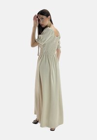 Abito maxi beige con corsetto arricciato, maniche a sbuffo e schiena scoperta. Tessuto leggero con una silhouette fluida e spacco laterale, abbinato a sandali neri.