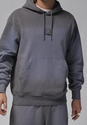 Mężczyzna w szarej bluzie z kapturem ze sznurkiem i kieszenią kangurka oraz naszywką z logo Air Jordan, w połączeniu z dopasowanymi szarymi dresami.