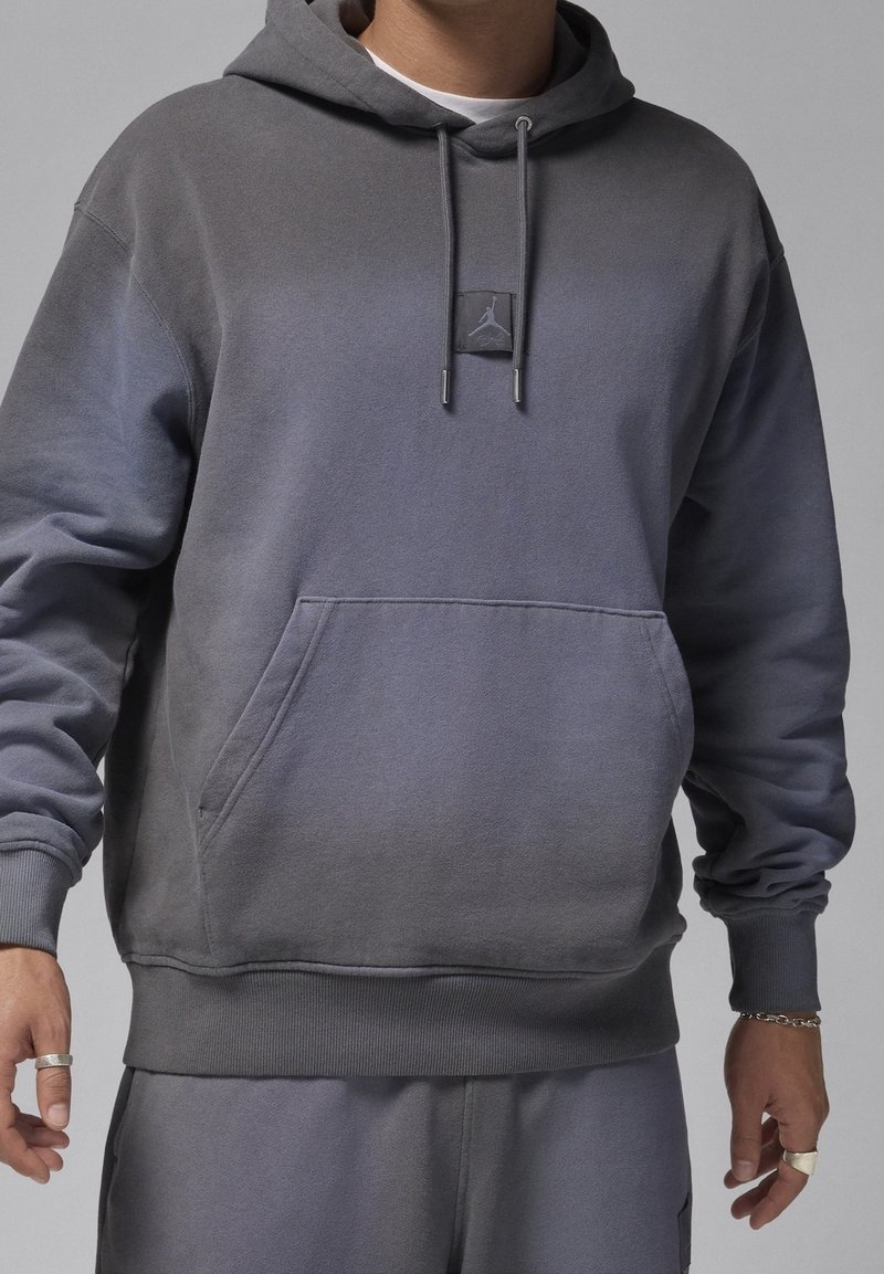 Homme portant un sweat à capuche gris avec cordon de serrage, poche kangourou et écusson du logo Air Jordan, assorti à un pantalon de survêtement gris.