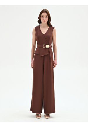 Donna in piedi, indossa una tuta marrone senza maniche con gambe larghe e una cintura abbinata, abbinata a sandali con zeppa beige.