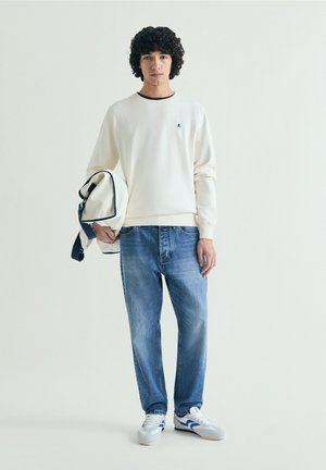 Jeune homme en sweat crème et jean bleu tient un sac à dos assorti crème et marine, debout devant un fond clair uni.