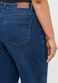 Blue denim jeans with a smooth texture, featuring a rear pocket, stitched seams, and a brown leather patch at the waistband.
